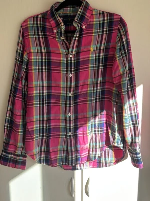Rutig skjorta från Ralph Lauren - Färgglad rutig skjorta från Ralph Lauren med klassisk button-down krage och lång ärm. Skjortan har en mix av rosa, blå, grön och vit i rutigt mönster samt den ikoniska broderade loggan på bröstet. Tillverkad i mjuk bomull och stängs med vita knappar framtill.