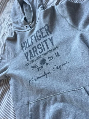 Grå Hilfiger Varsity hoodie - Snygg grå hoodie från Tommy Hilfiger med tryck 'Hilfiger Varsity' och Brooklyn Eagles på bröstet. Klassisk känguruficka, huva med dragsko och mjukt material. Perfekt för en avslappnad streetstyle-look.