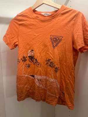 Orange t-shirt med cykeltryck från H&M - Cool orange t-shirt från H&M med tryck av en cyklist och palmer samt en liten logga på bröstet. Perfekt för dig som gillar streetstyle och sportiga vibes. Skön passform och tillverkad i mjuk bomull.