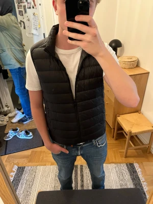 Uniqlo väst  - Snygg svart dunväst med ståkrage och quiltad design. Västen har dragkedja framtill och en stilren, minimalistisk look som passar perfekt till jeans och t-shirt. Perfekt nu under våren, mycket bra skick och inga defekter alls 