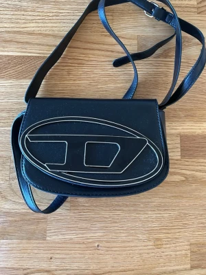 Svart axelväska från Diesel - Cool svart axelväska i slätt läder från Diesel med justerbar rem och ett stort, stilrent logomönster på framsidan. Väskan har en rundad form och stängs med lock. Perfekt för dig som gillar unik design och vill sticka ut.