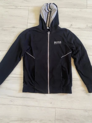 Original Hugo Boss zip hoodie -   Stilren hoodie från Hugo Boss i klassiskt svart utförande. Perfekt balans mellan sportig och elegant – lika snygg till jeans som till en mer uppklädd casual look.  ✨ Diskret broderad logga på bröstet ✨ Kontrastfärgad dragkedja och detaljer ✨ Mjuk och bekväm kvalitet ✨ Huvan har snygg logodetalj på insidan 
