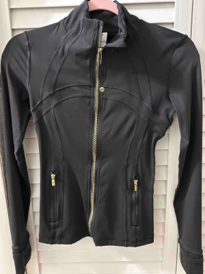 Svart jacka från Lululemon med guld-detaljer - Snygg svart jacka från Lululemon med figurnära passform och guldiga dragkedjor. Jackan har hög krage, långa ärmar och två fickor med dragkedja. Materialet är stretchigt och bekvämt, perfekt för en aktiv livsstil.