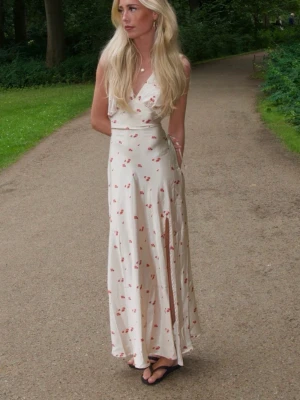 Lång blommig omlottklänning - Säljer en elegant långklänning i ljus beige med små röda blommor. Klänningen har omlott-design, v-ringning och är ärmlös. Materialet är troligtvis silke eller satin, vilket ger ett fint fall och glans. Perfekt för sommarens alla tillfällen.