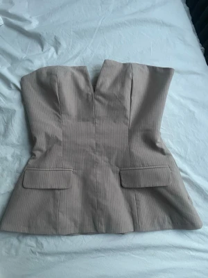 Beige randig korsettopp - Snygg beige korsettopp med diskreta vita ränder och markerad v-ringning. Toppen är axelbandslös och har två dekorativa fickor framtill. Perfekt för dig som vill ha en chic och stilren look.