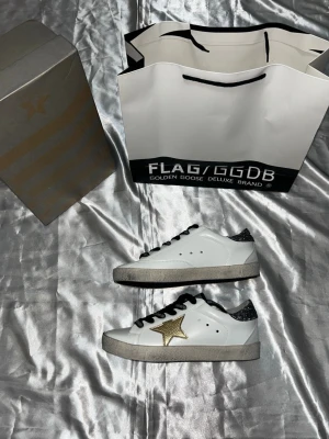 Golden Goose vita sneakers med stjärna - Säljer ett par vita Golden Goose sneakers med svartglittrande häl och svart snörning. Skorna har en ikonisk guldstjärna på sidan och en slitstark sula. Klassisk låg modell i läder som ger en cool och trendig vibe.24,5cm 