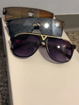 Louis Vuitton Solglasögon - Tre par Louis Vuitton solglasögon. Ett par med svart båge, guldiga detaljer och lila tonade glas, samt två olika par med monogrammönstrade sköldformade glas i brunt och grått. Båda har exklusiv design och tydliga LV-detaljer.