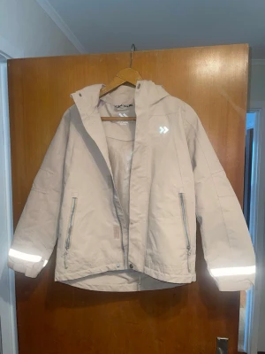 Beige vindjacka med reflex från Lager157 - Snygg beige vindjacka från Lager157 med reflexdetaljer på ärmar, rygg och bröst. Jackan har huva, två dragkedjeförsedda fickor och meshfoder på insidan. Perfekt för att hålla sig synlig och skyddad mot vind. Stängs med dragkedja och tryckknappar.