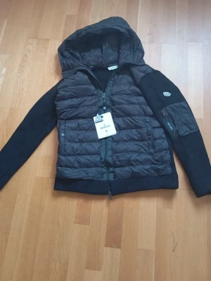 Svart cardigan Moncler - Svart cardigan från Moncler med vadderad front och huva i glansigt material samt stickade ärmar. Dragkedja framtill och ficka med dragkedja på ena ärmen. Klassisk Moncler-logga på ärmen. Perfekt för kyliga dagar. Sprillans ny. PRIS GÅR ATT FÖRHANDLA