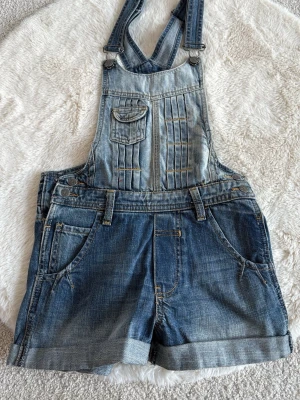 Jeansshorts byxdress med hängslen - Cool jeansbyxdress med shorts och justerbara hängslen. Den har fickor fram och bak, en liten bröstficka med lock och snygga sömdetaljer. Materialet är klassisk denim i blå nyanser med slitningar och uppvikta benslut. Perfekt för en avslappnad och trendig look.