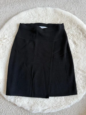 Svart kjol från Upgrade (S) - Svart stickad kjol från Upgrade i storlek S. Kjolen har en enkel och stilren design med hög midja och en diskret omlott-effekt framtill. Perfekt att matcha med en snygg topp eller hoodie för en cool look.