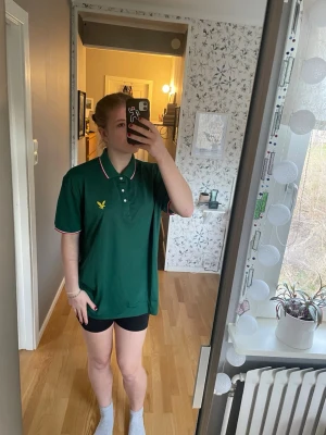 Grön pikétröja från Lyle & Scott - Snygg grön pikétröja från Lyle & Scott med klassisk krage och knappar framtill. Tröjan har vita och röda detaljer på krage och ärmslut samt den gula loggan över bröstet. 