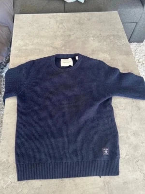 Mörkblå stickad tröja från GANT - Snygg mörkblå stickad tröja från GANT med rund halsringning och ribbade muddar vid ärmslut och nederkant. Tröjan har en diskret patch med GANT-logga nedtill och är tillverkad i ett grovt stickat material som ger en klassisk look.