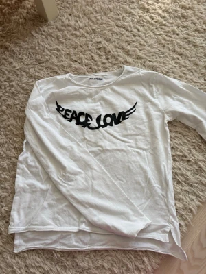 t-shirt Zadig & Voltaire - Säljer en vit långärmad t-shirt från Zadig & Voltaire med svart tryck 'PEACE LOVE' i cool design på bröstet. Tillverkad i mjuk bomull och har en avslappnad passform. Perfekt till jeans eller kjol för en chill look.