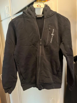Svart hoodie från Sail Racing - Svart hoodie från Sail Racing med dragkedja framtill och en extra bröstficka med dragkedja. Snygg logga på bröstet och huva. Perfekt för en chill och sportig stil.