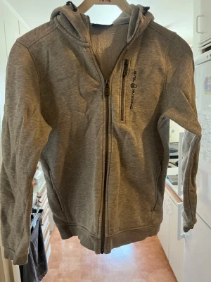 Grå zip hoodie Sail Racing Storlek 160 - Snygg grå hoodie från Sail Racing med dragkedja framtill och en bröstficka med zip. Mjuk fleece på insidan och broderad logga på bröstet samt på huvan. Perfekt för chill dagar och enkel att matcha med jeans eller joggers. Storlek 160.