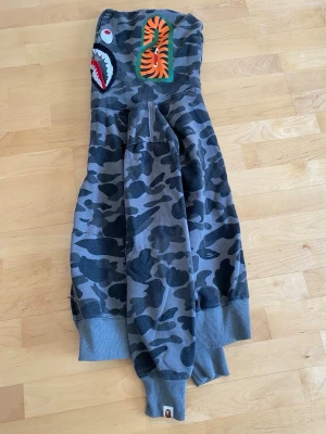 BAPE camo hoodie med haj och tiger - Säljer en grå camo hoodie från BAPE med ikoniska haj- och tigerpatches på huvan samt färgglada bokstäver. Hoodien har ribbade muddar och är tillverkad i mjuk bomullsblandning. Perfekt för dig som gillar streetwear och vill sticka ut. Storlek M