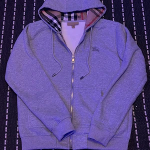 Grå hoodie från Burberry med dragkedja - Snygg grå hoodie från Burberry med klassiskt rutigt foder i huvan och broderad logga på bröstet. Tröjan har snörning i huvan, ribbade muddar och praktisk dragkedja framtill. Perfekt för dig som gillar stilrena och exklusiva plagg.