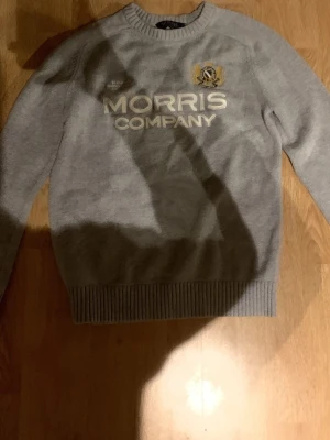 Grå stickad tröja Morris Company - Snygg grå stickad tröja från Morris Company med broderad logga och vit text framtill. Tröjan har rund hals, ribbade muddar vid ärmslut och nederkant samt lång ärm. Perfekt för dig som gillar klassisk stil med en modern touch. Köpt för ca 1100 kr o är använd ett fåtal gånger