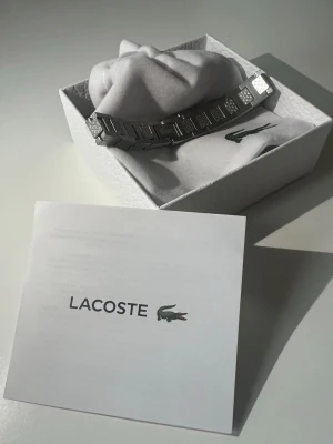 Silverfärgat armband från Lacoste - Snyggt armband i silverfärgad metall från Lacoste. Armbandet har en stilren design med rektangulära länkar och diskreta mönsterdetaljer på vissa länkar. Kommer i original ask med Lacoste-logga. Perfekt accessoar för dig som gillar minimalistisk och modern stil.