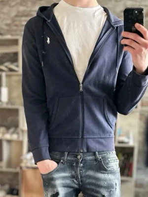 Mörkblå hoodie från Polo Ralph Lauren - Snygg mörkblå hoodie med dragkedja från Polo Ralph Lauren. Klassisk design med vit logga på bröstet, huva med dragsko och två fickor framtill. Tillverkad i mjukt bomullsmaterial som känns skönt mot huden. Perfekt för en avslappnad och stilren look. Modellen är cirka 180 o vägar 62 kg 
