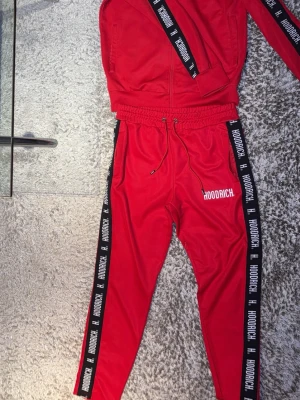 Rött Hoodrich loungewear set - Säljer ett rött loungewear set från Hoodrich med matchande ziptröja och byxor. Setet har svarta detaljer med Hoodrich-logga längs ärmar och ben, samt vita tryck på bröst och lår. Materialet är mjukt och stretchigt, perfekt för chill dagar hemma.
