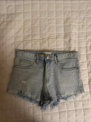 Ljusblåa högmidjade jeansshorts - Säljer ett par ljusblåa högmidjade jeansshorts med fransig kant och slitningar. Klassisk femficksmodell med knapp och dragkedja framtill. Perfekta för varma dagar och en avslappnad stil. Helt nya utan etikett. Säljs för att dem var för små och kunde inte returneras. Priset kan diskuteras 