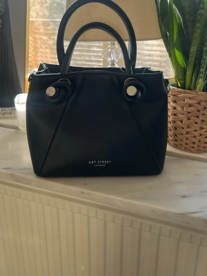 Svart handväska från Art Street - Snygg svart handväska från Art Street London med två handtag och dekorativa silverfärgade detaljer. Väskan har en stilren, fyrkantig form och är tillverkad i slätt konstläder. Praktisk dragkedja baktill och minimalistisk design som passar till många outfits.