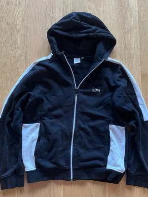 Svart hoodie från Hugo Boss - Säljer en svart hoodie från Hugo Boss med vit dragkedja och vita detaljer längs ärmarna. Hoodien har huva, fickor och logotypen BOSS Hugo Boss på bröstet. Perfekt för dig som vill ha en stilren och bekväm hoodie med exklusiv känsla.