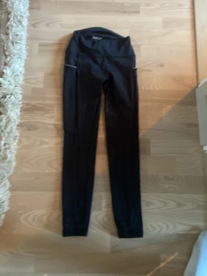 Svarta leggings från Röhnisch - Säljer ett par svarta leggings från  Horze med hög midja och reflekterande detaljer. De har en tight passform och är tillverkade i stretchigt funktionsmaterial, perfekta för ridning eller träning . Snygg logga baktill och diskreta mönster längs benen.