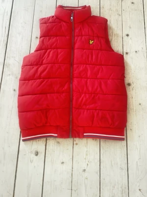Röd dunväst från Lyle & Scott - Snygg röd dunväst från Lyle & Scott med gul logga på bröstet. Västen har ståkrage, dragkedja framtill och ribbade muddar med vita och blå detaljer längst ner. Perfekt för lager på lager och ger en sportig look.