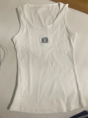 Vit ribbad linnetopp från Loewe - Vit ribbad linnetopp från Loewe med broderad svart logga framtill. Toppen är ärmlös och har en klassisk rund halsringning. Perfekt att styla med jeans eller kjol för en clean look.