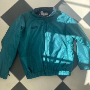 Turkos vindjacka från Columbia - Snygg turkos vindjacka från Columbia med dragkedja framtill, två fickor med dragkedja och resår vid ärmslut och midja. Jackan har en klassisk retrostil och står emot vind bra. Perfekt för dig som gillar streetwear och Gorpcore. Perfekt nu till sommaren och våren 