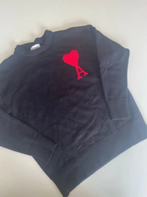 Svart stickad tröja med rött hjärta - Svart stickad tröja med rund hals och ribbade muddar. På bröstet finns ett rött hjärta och bokstaven A broderat. Perfekt för dig som gillar stilrena plagg med en twist. Tröjan har lång ärm och klassisk passform.