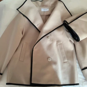 Beige kappa med svarta detaljer - Elegant beige kappa från Bubbelroom med svarta kanter och dubbelknäppning. Den är i storlek xs men funkar som storlek S också. Jackan är sparsamt använd💕