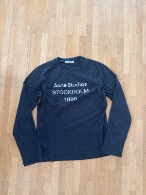 Acne Studios Long Sleeve  - Cool svart långärmad topp från Acne Studios med vit text 'Acne Studios STOCKHOLM 1996' på bröstet. Klassisk rund halsringning och relaxed fit. Perfekt till jeans eller kjol för en clean och trendig look.