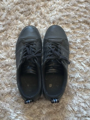 Svarta PS Paul Smith sneakers - Stilrena svarta sneakers från PS Paul Smith i läder med svarta skosnören och diskret logga på sidan. Låg modell med klassisk siluett och randig detalj på hälen. Perfekta för dig som gillar minimalistisk och clean stil.