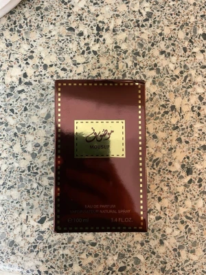 Mousuf Eau de Parfum 100 ml - Säljer en lyxig parfym från Mousuf, Eau de Parfum i 100 ml flaska. Förpackningen är mörkröd med gulddetaljer och har en elegant, fyrkantig form. Parfymen har orientaliska inslag och kommer i en snygg kartong med guldram och arabiska tecken.