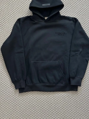 Svart Essentials hoodie Fear of God - Säljer en svart hoodie från Essentials Fear of God med diskret logga på bröstet och stor text på ryggen. Hoodien har en stor huva, ribbade muddar och en klassisk magficka. Materialet är mjukt och bekvämt, perfekt för en avslappnad look.