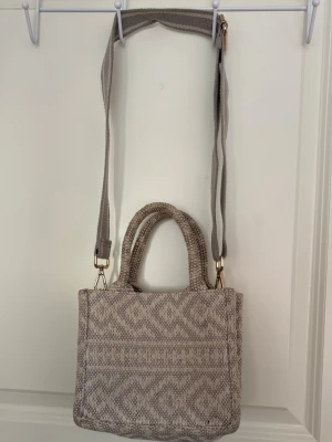 Beige mönstrad handväska med axelrem - Snygg beige handväska med stickat mönster och avtagbar, justerbar axelrem. Väskan har två handtag och ett diskret geometriskt mönster i ljusa toner. Perfekt för dig som gillar en unik accessoar med boho-vibe.