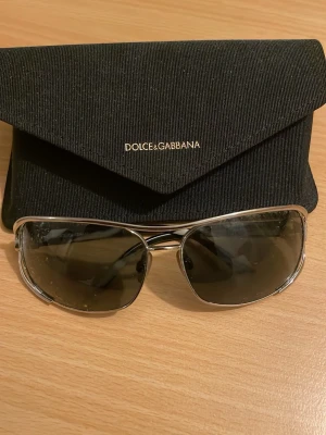 Dolce & Gabbana silver solglasögon - Snygga solglasögon från Dolce & Gabbana med silverfärgad metallram och mörka linser. Glasögonen har en klassisk aviatorform och kommer med ett svart fodral med logotyp i guld. Perfekt för dig som vill ha en stilren och exklusiv look.