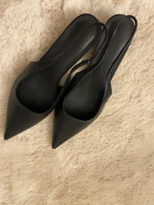 Svarta spetsiga pumps med slingback - Säljer ett par stilrena svarta slingback-skor med låg klack från nelly, Perfekta både till vardag och fest!  Skorna är helt oanvända och i nyskick – inga märken eller slitage. Köptes nyligen men har tyvärr inte kommit till användning.  * Storlek: 38 * Färg: Svart * Modell: Slingback med låg klack * Skick: Oanvända / nyskick  Enkla att matcha och väldigt bekväma tack vare den lägre klacken.