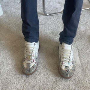 Maison Margiela paint splash sneakers - Maison Margiela sneakers i vitt läder och mocka med färgstänk. Klassisk låg modell med vita snören och brun sula. Snygg och unik design som sticker ut med sin konstnärliga touch. Box medföljer och man kan se lite skada vid sulorna på skorna. 