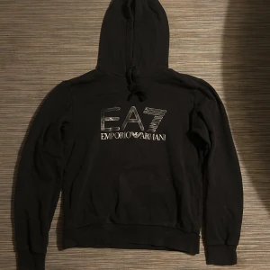 Svart EA7 hoodie från Emporio Armani - Svart hoodie från Emporio Armani EA7 med stor logga i silver på bröstet. Tröjan har huva med snörning och långa ärmar.  Inte använd så mkt. Pris går att diskutera 