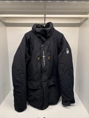 Sail racing jacka  - Svart dunjacka från Sail Racing med hög krage, två bröstfickor med gula dragkedjor och logga på ärmen samt ryggen. Jackan har justerbara ärmslut med kardborre och sportig design. Perfekt för kalla vinterdagar när du vill hålla värmen.
