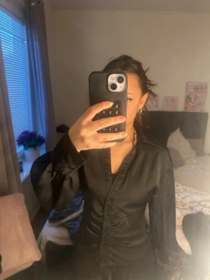 Svart satinblus med knappar - Snygg svart blus i satin med dekorativa knappar framtill och klassisk krage. Blusen har långa ärmar med breda manschetter och en figurnära passform som ger en elegant look. Perfekt för dig som gillar stilrena och trendiga plagg. Köparen står för frakt 