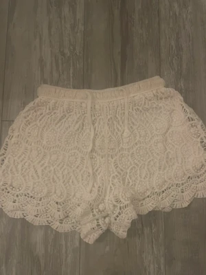 Vita virkade shorts med snörning - Säljer ett par vita virkade shorts med snygg snörning i midjan och vågig spetskant. De har ett detaljerat spetsmönster och är perfekta för sommaren. Köptes i Thailand! (Storlek står inte men skulle säga s)💓