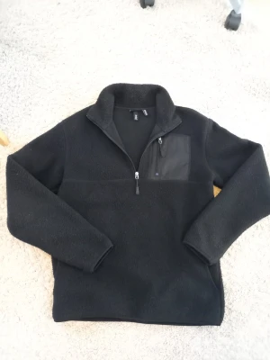 Svart fleece från H&M Thermolite - Svart fleece med relaxed fit från H&M, tillverkad i Thermolite-material. Tröjan har halv dragkedja framtill och en bröstficka med dragkedja. Perfekt för lager-på-lager och håller dig varm under kyliga dagar.
