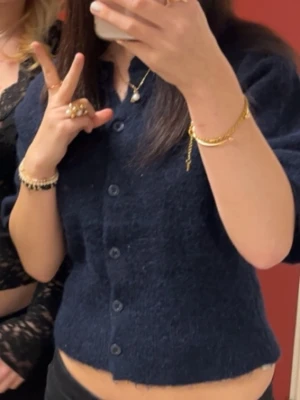 Mörkblå kortärmad kofta - Mysig mörkblå kofta med korta ärmar och knappar framtill. Koftan har en mjuk, lite luddig textur och en enkel, rak passform. Perfekt att styla över ett linne eller t-shirt för en avslappnad look.