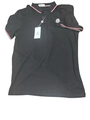 Svart Moncler pikétröja med logga - Klassisk svart pikétröja från Moncler med vit och röd rand på krage och ärmslut. Tröjan har korta ärmar och en broderad Moncler-logga på bröstet. Tillverkad i bomull för en bekväm och stilren look.